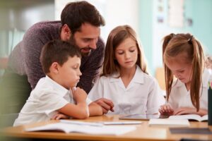 Male Teacher With Three Elementary School Pupils Wearing Uniform Using Digital Tablet At Desk ways to test student learning